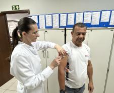 #ParaQuemNaoPodeVer Cena dentro de um consultório ou ambiente de saúde. Uma mulher de cabelo preso, óculos e jaleco branco está aplicando uma injeção no braço de um homem. O homem veste uma camiseta branca com a palavra "ENFERMAGEM" estampada. Eles estão em pé, próximos a armários brancos, com vários papéis presos em pastas azuis na parede ao fundo. Há também uma placa de saída de emergência verde na parede superior direita.