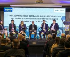 #ParaQuemNaoPodeVer Foto de um painel em um evento corporativo, com cinco pessoas (quatro homens e uma mulher) sentadas no palco, participando de um debate. No fundo, há um grande telão com o texto: "Logística no Brasil ONDE ESTAMOS HOJE E COMO ALCANÇAR OS INVESTIMENTOS NECESSÁRIOS PARA O DESENVOLVIMENTO ECONÔMICO SUSTENTÁVEL DA REGIÃO SUL". O público está sentado de frente para o palco, acompanhando a discussão. No canto inferior direito da tela, há um púlpito transparente com a mesma identidade visual do 