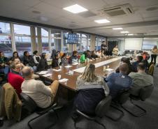 #ParaQuemNaoPodeVer Foto mostra uma reunião em uma sala de conferências. Ao redor de uma mesa grande e retangular, há muitas pessoas sentadas, a maioria prestando atenção em alguém que está falando ou apresentando (próximo a um quadro de projeção na ponta da mesa). A sala tem janelas grandes ao fundo, por onde é possível ver navios. As pessoas na reunião são de perfis variados e algumas estão com laptops ou papéis na mesa. A imagem sugere um ambiente corporativo ou de trabalho colaborativo. 