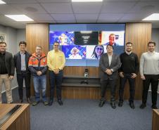 #ParaQuemNaoPodeVer Foto de sete homens em uma sala de reunião corporativa. Eles estão todos em pé, lado a lado, olhando para a câmera. Quatro deles estão vestidos de forma casual e três usam roupas mais formais (terno ou camisa social). Dois dos homens ao centro estão utilizando uniformes: um de jaqueta laranja e outro de jaqueta amarela, que parecem ser de alguma empresa ou serviço. Atrás deles, há uma televisão grande na parede exibindo uma imagem colorida com pessoas em uma chamada de vídeo, sugerindo q