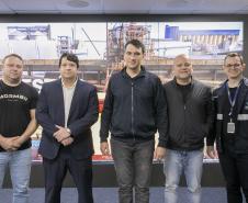 #ParaQuemNaoPode Ver Imagem mostra cinco homens posando para uma foto em grupo. Eles estão em pé, lado a lado, com um fundo que parece ser uma imagem de uma área industrial ou de construção, com estruturas metálicas e guindastes. Um dos homens, na parte superior esquerda, veste uma camiseta preta com a palavra "Mormaii" escrita. Outro homem, ao lado dele, está de terno azul escuro. Os demais usam roupas casuais ou jaquetas, e um deles, na parte inferior, usa um crachá e uniforme preto com detalhes 