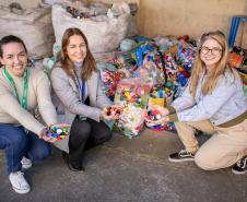 #ParaQuemNaoPodeVer Foto de três mulheres sorrindo, agachadas ou ajoelhadas próximas umas das outras. Elas estão segurando várias tampinhas plásticas coloridas nas mãos. Ao fundo, há vários sacos grandes cheios de tampinhas plásticas, sugerindo uma campanha de reciclagem ou coleta de tampas. O ambiente parece ser um galpão ou área de armazenamento. As três estão vestidas de forma casual e aparentam estar felizes com a atividade.
