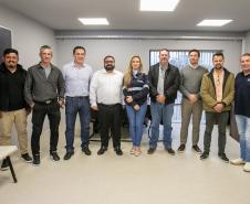 #ParaQuemNaoPodeVer Foto mostra um grupo de nove pessoas, todas em pé e organizadas em fila. O ambiente parece um escritório, com paredes claras, piso claro e janelas atrás do grupo. As pessoas estão vestidas de maneira casual a social, com camisas, jaquetas, calças jeans e algumas de camiseta. Há uma mulher no grupo enquanto o restante do grupo é composto por homens. Todos posam para a foto sorrindo. 