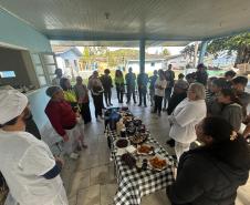 #praquemnaopodever foto mostra um grupo de pessoas em círculo em volta de uma mesa com toalha xadrez e repleta de comida. A maioria olha para uma pessoa que fala no outro lado da foto.