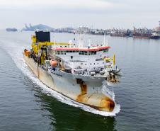 #ParaQuemNaoPodeVer Foto de um navio grande navegando em um corpo d'água, provavelmente próximo a um porto. O navio está indo em direção à direita da imagem e deixando um rastro na água. O navio é branco com detalhes amarelos e laranja, e parece equipado para operações industriais ou de transporte pesado, com guindastes e equipamentos grandes na parte traseira. Ao fundo, vê-se uma área portuária com guindastes, navios atracados e estruturas industriais próximas à água.