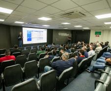#ParaQuemNaoPodeVer Auditório com diversas pessoas sentadas em cadeiras assistindo a uma apresentação. Na frente, um homem está em pé próximo a um telão, onde uma apresentação de slides é projetada. Atrás dele, há bandeiras do Brasil e de outro estado ou instituição. O ambiente tem iluminação de teto forte, ar-condicionado e paredes claras. As pessoas parecem atentas à apresentação. A tela mostra informações em português, porém está levemente desfocada na imagem.
