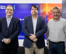 #ParaQuemNaoPodeVer Foto de três homens posando para a câmera. Eles estão de pé lado a lado. Dois deles usam ternos com camisa clara, e um, na frente, está sem paletó, usando uma camisa social listrada clara e jeans. Eles sorriem levemente. Ao fundo, aparece um painel iluminado com cores azuis e laranja, com parte da palavra “Gartner” visível.
