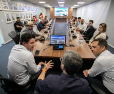 #praquemnaopodever foto mostra um homem sentado de costas para a camera, na ponta de uma mesa em formato de u e rodeado de pessoas sentadas com roupas formais olhando para ele.