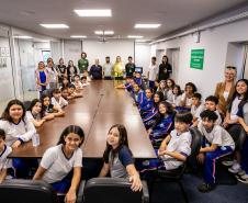 #ParaQuemNaoPodeVer Foto mostrando um grupo grande de pessoas sentadas ao redor de uma mesa de reunião em uma sala fechada. A maioria aparenta ser crianças e adolescentes, algumas usando uniformes escolares de calça azul e camiseta branca, enquanto adultos estão espalhados entre eles, talvez professores ou responsáveis. Todos olham para a câmera e sorriem, parecendo participarem de algum evento escolar ou visita guiada. No fundo, há quadros na parede e uma porta de vidro; do lado direito, há avisos e um car
