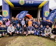 #ParaQuemNaoPodeVer Foto de um grupo grande de pessoas posando para a câmera, todas sorrindo, em frente a banners e bandeiras com os logotipos da Polícia Rodoviária Federal (PRF) e “Portos do Paraná – Logística. Eficiência. Valor.” Ao fundo, há balões laranja reunidos decorando a entrada de um prédio. As pessoas estão ao ar livre sobre uma área gramada, usam roupas variadas, incluindo uniformes da PRF, coletes e agasalhos. O ambiente é de evento institucional ou comemorativo, possivelmente uma ação conjunta