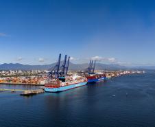 #ParaQuemNaoPodeVer Foto aérea de um porto. Na imagem, há um navio cargueiro grande, de cor azul, atracado em um píer. Próximo ao navio, vê-se diversos contêineres coloridos e guindastes altos, típicos de portos de carga. O mar ao redor é azul e há uma cidade ao fundo, com vários prédios e montanhas ao longe. A imagem está levemente inclinada para a direita.