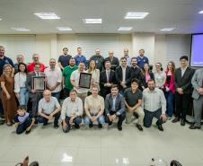 #ParaQuemNaoPodeVer Foto de um grupo grande de pessoas em um ambiente fechado, possivelmente uma sala de reuniões ou auditório. Todos estão vestidos de maneira formal e informal, sugerindo um evento corporativo ou de premiação. Algumas pessoas seguram placas ou certificados, indicando que receberam algum tipo de reconhecimento. Ao fundo, há bandeiras, reforçando o clima institucional do local. As pessoas estão sorrindo e posando para a foto, aparentando clima de celebração ou comemoração.