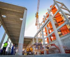 #praquemnaopodever foto do chão do canteiro de obras do Moegão, com os pilares cinzas e máquinas laranjas, trabalhadores estão no local espalhados trabalhando e o céu está de fundo, em um azul claro.