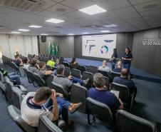 #ParaQuemNaoPodeVer Sala de apresentação com várias pessoas sentadas em cadeiras, assistindo a uma palestra. Na frente, há um telão exibindo gráficos e textos, além de duas pessoas apresentando. No fundo, há bandeiras do Brasil e do Paraná, e na parede está escrito "Portos do Paraná". O ambiente é bem iluminado, com teto branco e piso azul. A atenção das pessoas está voltada para o telão e os palestrantes.