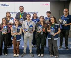 #ParaQuemNaoPodeVer Foto de um grupo de dez pessoas em pé, posando para a câmera em um ambiente interno, possivelmente em um evento ou cerimônia. Todas parecem estar sorrindo. Algumas seguram troféus e brindes, sugerindo premiação ou reconhecimento. O fundo tem um painel branco com texto, onde é possível ler “PORTOS DO PARANÁ”, “programa”, “digitalização”, e “HACK3R”. A maioria das pessoas usa crachás e roupas casuais ou uniformes. 