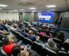#ParaQuemNaoPodeVer Imagem de uma sala de conferências. Na foto, várias pessoas estão sentadas em poltronas, formando fileiras, e assistindo a uma apresentação feita por duas pessoas na frente da sala, próximo a um telão. No telão, há um slide azul com informações (textos e gráficos) e, na parede, o logotipo “Portos do Paraná” está visível. Também estão presentes bandeiras do Brasil.