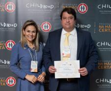 #ParaQuemNaoPodeVer Foto de um homem e uma mulher sorrindo, posando juntos para a câmera. Eles estão em frente a um painel de fundo decorado com os logos do "Programa Na Mão Certa" e da "Childhood Brasil", ambos destinados à proteção da infância. O homem está com um paletó azul, camisa branca e gravata amarela, segurando um certificado ou diploma. A mulher está usando uma roupa social azul claro, com crachá pendurado no pescoço. Ambos parecem estar participando de um evento formal ou solenidade de premiação