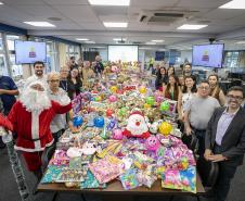 #praquemnaopodever a foto mostra uma mesa de madeira comprida e com quase mil brinquedos em cima de todas as cores e formatos. Nas lateriais, dezenas de pessoas estão a postos sorrindo e posando para o fotógrafo dentro de uma sala de reunião com tapete cinza e janelas grandes