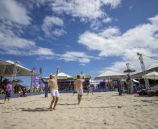 #praquemnaopodever imagem na praia, dentro da quadra de areia, a dupla de homens de bermuda branca e camisa amarela estão a frente de costas pra foto. Do outro lado está a outra dupla pronta pra receber a bola. O céu está azul com algumas nuvens. Ao fundo e dos lados as arquibancadas estão lotadas