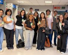 #ParaQuemNaoPodeVer Grupo de onze jovens alinhados em um corredor interno, todos segurando crachás verdes voltados para a frente. Na foto, as pessoas estão em pé, lado a lado, sorrindo. Há diversidade de tons de pele, de claros a médios e mais escuros, e estilos de cabelo diferentes, incluindo cabelos lisos, cacheados e ondulados, alguns longos, outros curtos, em cores como castanho, preto e loiro. As roupas são casuais: calças jeans claras e escuras, calça de moletom cinza, calça preta, tênis brancos