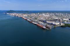 A imagem mostra o Porto de Paranaguá visto do alto, com uma grande faixa da Baía de Paranaguá na parte de baixo da foto até o final do porto. Os navios estão pequenos e enfileirados, a maioria na cor vermelha.