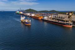 #paraquemnaove Imagem mostra grande parte do mar azul à esquerda, e à direita está o Porto de Paranaguá com navios atracados por todo o cais. No mar, segue navegando um navio vermelho de com faixa azul. O céu está aberto, com poucas nuvens.
