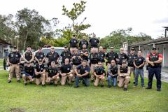 Dezenas de guardas portuárias estão fardados e posando para a foto lado a lado em um gramado. A maioria veste uma calça creme com camisa preta e colete, com os braços cruzados à frente.
