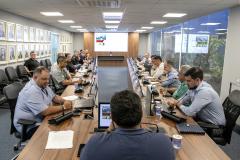 #ParaQuemNaoPodever Na foto, há várias pessoas sentadas ao redor de uma longa mesa de conferência. As pessoas estão usando roupas formais e parecem estar participando de uma reunião ou discussão. Há microfones na mesa em frente a cada pessoa, e algumas delas estão usando laptops ou tablets. A sala tem uma parede de vidro ao fundo, através da qual é possível ver plantas. Na parede oposta, há quadros com retratos de pessoas. A iluminação é feita por lâmpadas embutidas no teto.