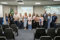 #ParaQuemNaoPodeVer A imagem mostra um grupo de 12 pessoas posando para uma foto em uma sala de conferências. Ao fundo, há duas bandeiras, uma do Brasil e outra de um estado ou município. Todos estão sorrindo e parecem estar participando de um evento ou reunião. No canto direito da imagem, há um mapa grande na parede. No canto superior direito, há um projetor exibindo uma imagem que parece