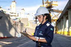 Portuária de uniforme azul de manga com o logo estampado "portos do parana" escreve em uma prancheta com a mão esquerda, enquanto um navio cinza ao fundo é carregado. Ela está de capacete branco e usa epis completos. #paraquemnaopodever