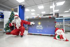 #ParaQuemNaoPodeVer A imagem mostra um homem vestido de Papai Noel agachado ao lado de uma árvore de Natal decorada com bolas azuis e douradas e uma estrela dourada no topo. Ele está usando um traje vermelho com detalhes brancos, incluindo um gorro e uma longa barba branca. O homem está fazendo um gesto de "paz e amor" com a mão direita. Ao lado dele, há um painel azul com a inscrição "Natal Solidário" em letras brancas e uma estrela amarela acima do texto. Na frente do painel, há um tapete ou almofada com 