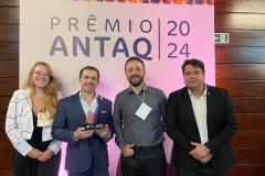 Quatro pessoas estão lado a lado, em frente a uma parede de madeira com um banner escrito "prêmio antaq 2024". São três homens e uma mulher com roupas formais, sorrindo e um deles segura um prêmio nas mãos. #praquemnaove