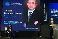 #ParaQuemNaoPodever A imagem mostra uma apresentação ou conferência. No centro da tela, há um slide com fundo azul escuro e texto em branco e azul claro. O texto diz "Dr. Luiz Fernando Garcia" e "Presidente da Portos do Paraná". Abaixo, está escrito "OS MAIORES NOMES DA ÁREA TRABALHISTA". No canto inferior direito do slide, há um logotipo circular azul com linhas horizontais.  Na parte inferior da tela, há uma foto de um homem de terno e gravata, que parece ser o Dr. Luiz Fe