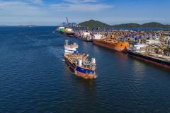 #ParaQuemNaoPodeVer A imagem mostra uma fotografia de um porto com vários barcos atracados. A água do mar é de um azul intenso e há uma grande quantidade de barcos coloridos, principalmente em tons de branco e azul, ao longo de um cais. Ao fundo, é possível ver uma linha de montanhas sob um céu azul claro.