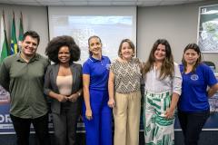 #ParaQuemNaoPodeVer A imagem mostra seis pessoas posando para uma foto em um ambiente interno. Há um homem de camisa verde e duas mulheres, uma com cabelo afro e outra com cabelo preso. Na fileira da frente, há três mulheres, uma usando uma camisa azul, outra com uma blusa estampada e a terceira com uma blusa branca e saia com listras verdes. Ao fundo, há bandeiras e um painel que parece ter um mapa ou uma imagem de cidade.