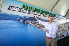 #paraquemnaopodever Homem de camisa banca, calça marrom e cinto marrom aponta para outdoor atrás com frase "R$ 1 Bilhão em investimentos"