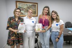 #ParaQuemNaoPodeVer A imagem mostra quatro pessoas posando para uma foto em um ambiente interno. Duas mulheres estão em pé na parte de trás, uma delas usando um vestido estampado colorido e óculos, segurando um papel, e a outra usando uma blusa branca. Na frente, há duas mulheres, uma usando uma blusa rosa e segurando um pacote, e a outra com uma camiseta branca com um desenho azul. Ao fundo, há uma pintura emoldurada na parede.