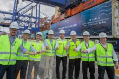 #paraquemnaopodever nove homens de coletes verdes e capacetes brancos seguram taças de champagne com os braços esticados para a frente. Ao fundo, guindastes azuis de mais de 50 metros de altura movimentam contêineres e à direita um outdoor em um contêiner com os dizeres "1,5 million TEU achieved"