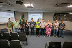 #ParaQuemNaoPodeVer A imagem um grupo de pessoas posando em um auditório. Há nove pessoas na foto, dispostas em duas fileiras. A fileira de trás tem quatro pessoas, e a fileira da frente tem cinco pessoas. No fundo, há uma bandeira do Brasil e um projetor de teto. As pessoas estão em pé em frente a uma tela de projeção que exibe um slide com texto, mas o texto não está totalmente legível na imagem. Algumas das pessoas estão segurando troféus ou prêmios. O ambiente parece ser uma sala de conferências ou um a