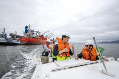 #praquemnaopodever quatro pessoas aparecem de colete laranja conversando dentro de uma lancha branca, que cruza a baía. Ao fundo, o céu é cinza e navios estão atracados no cais de Paranaguá. 