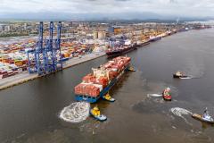 #ParaQuemNaoPodeVer A imagem mostra um grande navio cargueiro carregado com contêineres coloridos, que está sendo auxiliado por três rebocadores na água. O navio está próximo a um cais onde há uma grande quantidade de contêineres empilhados e guindastes azuis para movimentação de carga. Ao fundo, é possível ver uma área urbana com várias construções e, mais distante, montanhas.