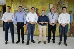 #ParaQuemNaoPodeVer A imagem mostra uma foto de seis homens em pé, posando para a câmera. Eles estão em duas fileiras, três na frente e três atrás. Todos estão vestidos de forma formal, com camisas sociais e calças. O fundo da foto é uma pintura mural colorida que parece representar uma cena cultural ou histórica.