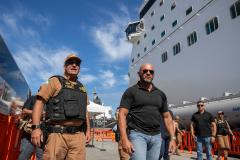 #ParaQuemNaoPodeVer A imagem mostra dois homens em destaque. O homem à esquerda está usando um uniforme marrom com um colete preto e um boné, além de óculos escuros. Ele parece ser um oficial de segurança. O homem à direita está vestindo uma camisa preta e também usa óculos escuros. Ao fundo, há um grande navio de cruzeiro branco, com várias janelas visíveis, e o céu está azul com algumas nuvens. 