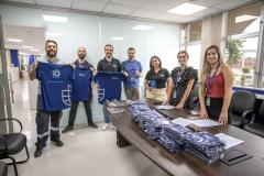 #ParaQuemNaoPodeVer A imagem mostra um grupo de pessoas em um ambiente de escritório. Na parte superior da imagem, há quatro homens em pé, segurando camisetas azuis com um logotipo branco. Eles estão sorrindo e parecem estar participando de algum tipo de evento ou celebração. Na parte inferior da imagem, há três mulheres e um homem em pé atrás de uma mesa, que está coberta com várias pilhas de camisetas azuis dobradas. As mulheres estão sorrindo e também parecem estar participando do mesmo evento.