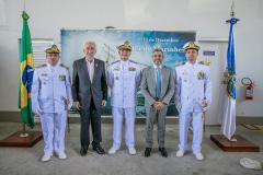 #ParaQuemNaoPodeVer A imagem mostra cinco homens posando para uma foto em frente a uma bandeira do Brasil e uma bandeira da Marinha do Brasil. Três deles estão vestidos com uniformes brancos de oficiais da Marinha, com chapéus brancos e insígnias no peito. Os outros dois homens estão usando ternos. Ao fundo, há um banner azul com o texto "13 de Dezembro" e "Dia do Marinheiro". A foto parece ter sido tirada em um ambiente interno, com uma parede branca ao fundo.