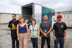 #praquemnaopodever cinco pessoas lado a lado posam para a foto em frente a um caminhão azul e verde que está com as portas da caçamba aberta e cheia de caixas com roupas.