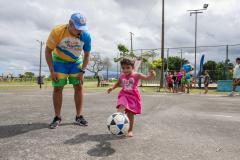 #praquemnaopodever uma criança de dois anos com um vestido rosa, chuta uma bola de futebol enquanto um representante do Governo do Paraná, com uma camisa colorida do verão maior, observa do lado esquerdo, encurvado, observando o chute da criança. ao fundo pessoas interagem no Aeroparque de Paranagua