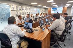#ParaQuemNaoPodeVer A imagem mostra uma foto de uma reunião em uma sala de conferências. Há várias pessoas sentadas ao redor de uma mesa de madeira, todas olhando para a frente, onde parece haver uma apresentação ou videoconferência em andamento. Na parede ao fundo, há várias fotos emolduradas. As pessoas estão usando roupas formais, como camisas sociais. Na mesa, há laptops, garrafas de água e outros materiais de escritório. A sala é bem iluminada, com luzes no teto.