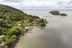 #ParaQuemNaoPodeVer A imagem mostra uma vista aérea de uma área costeira com vegetação densa e casas próximas à água. Há uma pequena península ou ilha coberta de árvores e algumas construções. A água ao redor é calma, e há algumas pequenas embarcações ou estruturas flutuantes próximas à costa. O céu está parcialmente nublado.