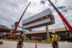 #ParaQuemNaoPodeVer A imagem mostra uma fotografia de um local de construção. Na foto, há dois trabalhadores usando equipamentos de segurança, como capacetes e coletes refletivos. Eles estão observando uma grande estrutura metálica sendo levantada por um guindaste vermelho. Ao fundo, há uma construção industrial com várias vigas e estruturas de aço. O céu está nublado.